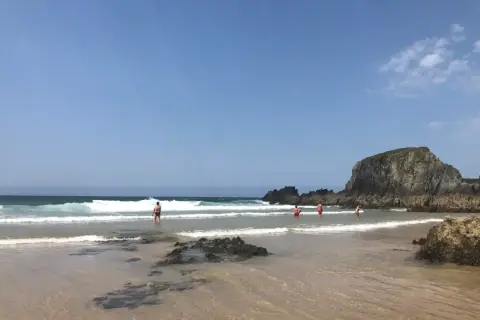 Gente disfrutando del mar en la playa de Frouxeira, con olas rompiendo y una gran roca verde al fondo bajo un cielo azul.