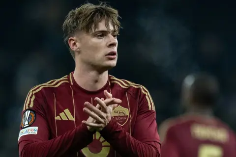 Piłkarz Nicola Zalewski w bordowej koszulce AS Roma z żółtymi pasami na ramionach.