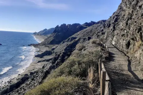 Sendero costero con vistas al mar Mediterráneo y montañas. Un camino de tierra con barandilla de madera serpentea por la ladera, ideal para un paseo marítimo en Mojácar Playa.
