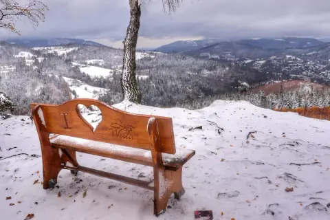 Drewniana ławka z wyciętym sercem i napisem "miasto Wisła" na tle zimowego krajobrazu. Widok na ośnieżone wzgórza i dolinę.