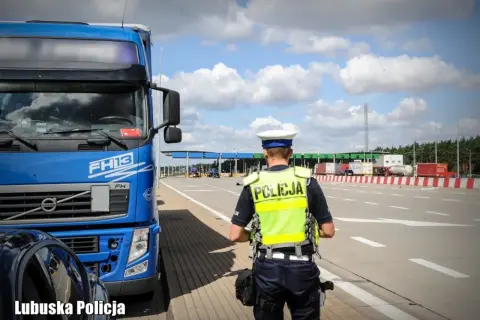 Policjant w kamizelce odblaskowej przy niebieskiej ciężarówce Volvo FH13. W tle przejście graniczne, kategoria c.