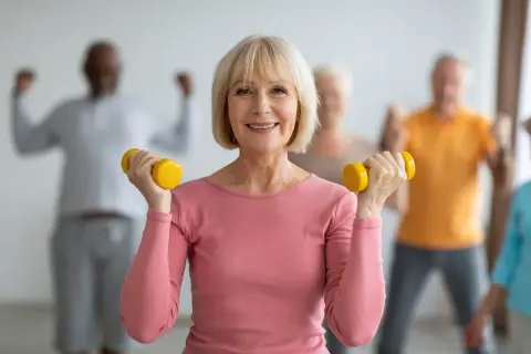 Une femme souriante fait de la gymnastique senior avec des haltères jaunes. D'autres personnes âgées s'entraînent en arrière-plan.