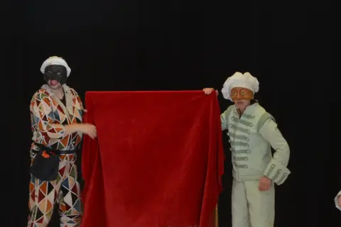 Dos actores con máscaras y trajes de época, uno con un traje de rombos y otro con un traje verde pálido, presentan una tela roja. Parecen parte de una obra de teatro de **els joglars zaragoza**.