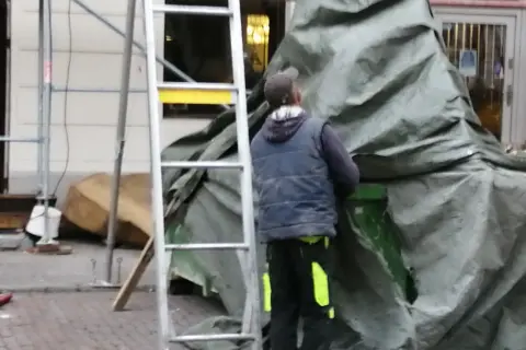 Pracownik w kamizelce i czapce przykrywa coś zieloną plandeką. Czy to hałas?