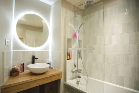 Salle de bain sans fenêtre, lumineuse avec un grand miroir rond éclairé, une vasque moderne et une douche à l'italienne.