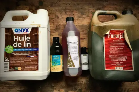 Plusieurs bouteilles d'huile pour le bois, dont de l'huile de lin et du goudron de pin, sont alignées sur une surface en bois.