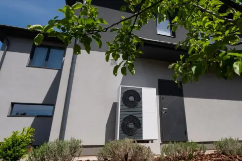Unità esterna di una pompa di calore residenziale, moderna e discreta, installata accanto a una porta d'ingresso su un edificio grigio.