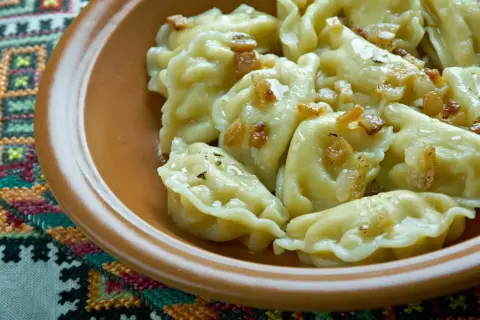 Pyszne pierogi ruskie z podsmażoną cebulką i skwarkami, podane w glinianej misce na haftowanej serwetce.