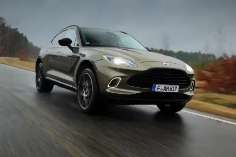 Elegancki Aston Martin SUV przemierza mokrą drogę wśród jesiennych drzew.
