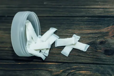 Białe woreczki z nikotyną, popularne jako snus zęby, rozsypane obok plastikowego pojemnika na drewnianym tle.