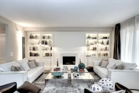 Soggiorno elegante con divani bianchi, librerie illuminate e camino. Le pareti color bianco latte creano un'atmosfera luminosa e ariosa.