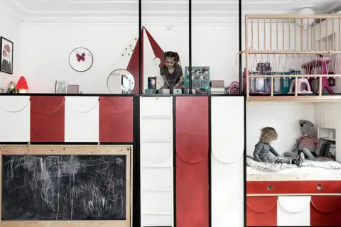 Zwei Kinder teilen sich ein Zimmer mit kreativen Schlafkojen und Spielbereichen. Ein Kind sitzt auf einem Bett, das andere spielt oben.