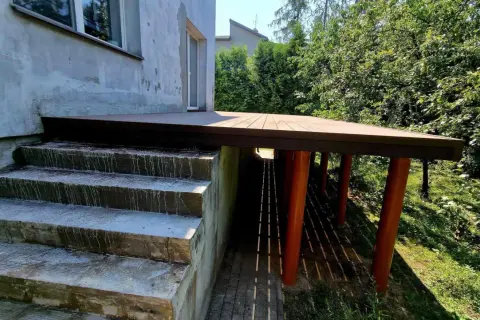 Nowy taras na słupach stalowych, z drewnianą podłogą, sąsiaduje z betonowymi schodami i zielonym ogrodem.