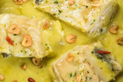 Merluza al pil pil, con trozos de pescado blanco en una salsa amarilla cremosa, adornada con láminas de ajo crujiente y guindilla.