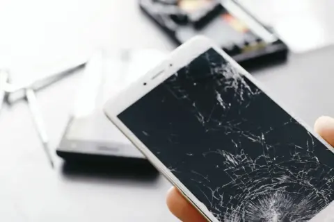 Dłoń trzyma telefon z pękniętym ekranem. Obok leżą narzędzia do naprawy.