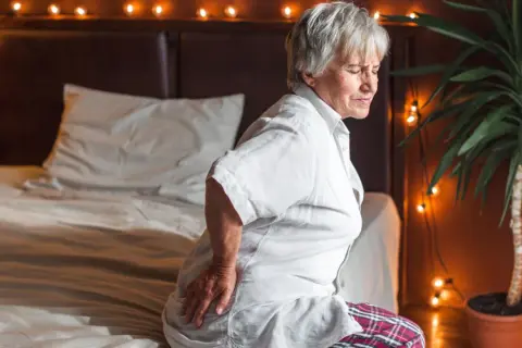 Elderly woman grimaces in pain, holding her hip. She looks like she slept on her hip wrong, sitting on a bed with festive lights.