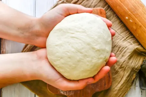 Dłonie trzymają kulę puszystego ciasta na pizzę włoską, gotowego do rozwałkowania. Obok wałek i lniana ściereczka.