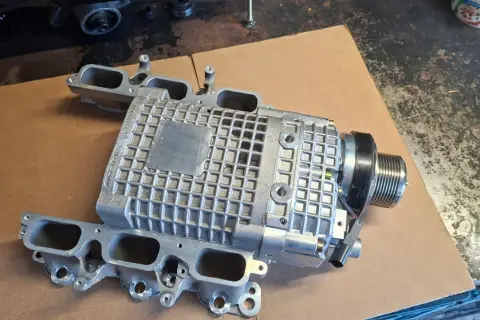 Srebrna sprężarka mechaniczna z kolektorem dolotowym, część do Mercedes Benz C32 AMG, leży na kartonie.