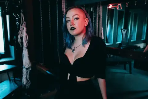 Eine Frau mit blauen Haaren und auffälligem Make-up steht in einem Raum mit Ketten und Seilen. Sie verkörpert die Frage: was ist ein Masochist?
