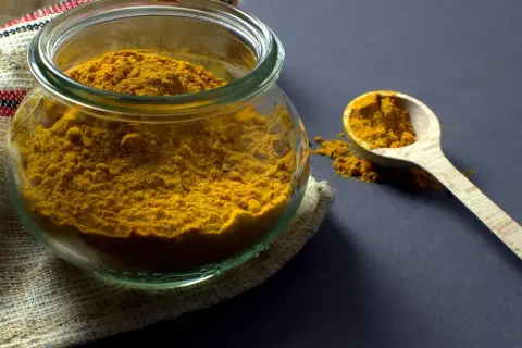 Un pot en verre rempli de poudre de curcuma, avec une cuillère en bois à côté. Ces épices, comme le cumin, apportent couleur et saveur.