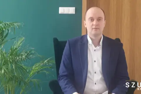 Mężczyzna w garniturze siedzi w fotelu obok rośliny, na tle zielonej ściany i drzwi.