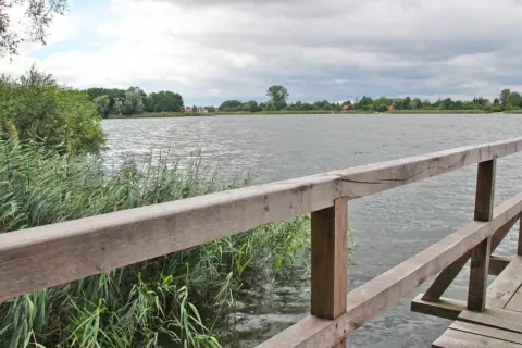 Drewniany pomost nad jeziorem, gdzie można łowić ryby. W oddali widać zalesiony brzeg i domy.