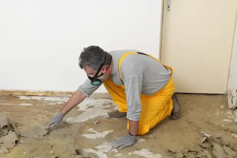 Un homme en salopette jaune et masque retire la colle de moquette avec une spatule. Une méthode naturelle pour enlever colle moquette.