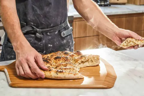 En person i förkläde håller upp en bit av en nybakad, ångande ramazan pidesi. Brödet är gyllene med sesamfrön.