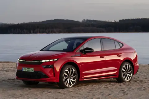 Czerwony Skoda Enyaq na piaszczystym brzegu, z lodowym jeziorem i lasem w tle.