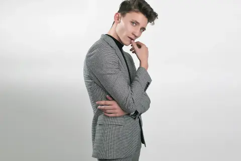 Elegancki outfit na 18 męski: młody mężczyzna w czarnym golfie i stylowej, kraciastym garniturze pozuje z dłonią przy ustach.