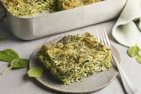 Eine Portion Spinat Ricotta Lasagne auf einem Teller, daneben eine Auflaufform mit der restlichen Lasagne. Frische Spinatblätter liegen daneben.
