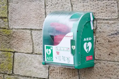 Boîtier vert fixé sur un mur de pierre, contenant un défibrillateur. Des pictogrammes indiquent qui peut utiliser un défibrillateur et comment l'utiliser.