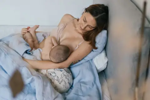 Matka w wygodnej pozycji biologicznej do karmienia, przytulona do poduszki, karmi piersią swoje dziecko.