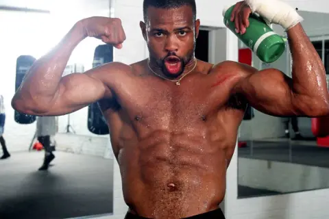 A sweaty boxer flexes his biceps in a gym, holding a green water bottle. He's training hard, ready for any division, perhaps even the Roy Jones Jr. division.