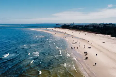 Plaża z ludźmi spacerującymi, morze i port z latarnią morską w tle.