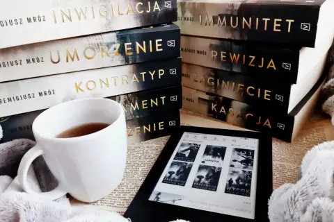 Stos książek Remigiusza Mroza, biały kubek z herbatą i czytnik Kindle.