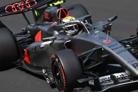 Un coche de **Audi F1** gris y negro con detalles rojos acelera en la pista. El piloto lleva casco amarillo y blanco.