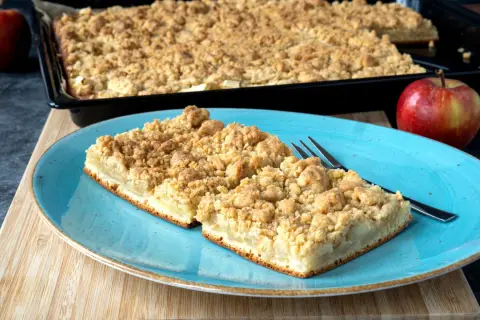 Zwei Stücke saftiger Apfelkuchen mit Streuseln auf einem blauen Teller, daneben ein ganzer Apfel. Perfekt für ein Apfelkuchen Rezept.