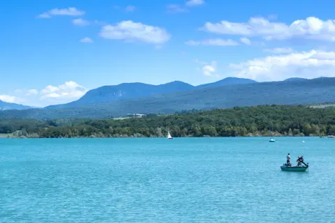 Le lac de Montbel, aux eaux turquoise, accueille un voilier et un petit bateau de pêche sous un ciel bleu parsemé de nuages.