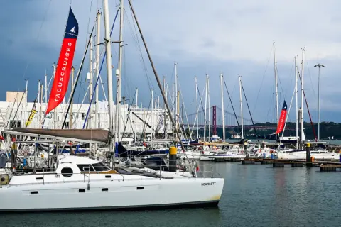Una **barca lunga e sottile**, la Scarlett, è ormeggiata in un porto pieno di altre barche a vela. Sullo sfondo, un ponte rosso attraversa il fiume.