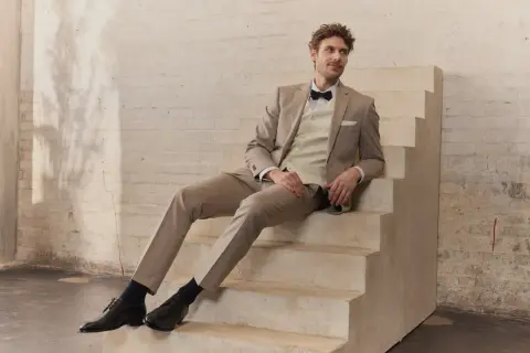 Mann in hellem Hochzeitsanzug mit Fliege sitzt auf Treppe. Perfekt für die Frage: wann Hochzeitsanzug kaufen?
