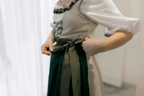 Eine Frau bindet die Schleife ihres Dirndls. Das Dirndl hat eine helle Weste mit Schnürung und eine dunkelgrüne Schürze.