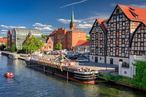 Malownicze stare miasta w Polsce nad rzeką. Barka z rzeźbą, łódka z ludźmi, zabytkowe kamienice i nowoczesna architektura.