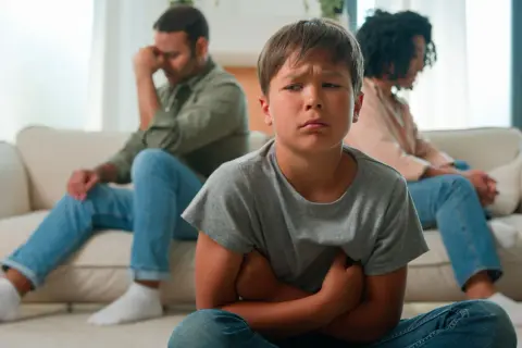 Bambino triste in primo piano, con i genitori separati seduti alle sue spalle, voltati l'uno dall'altra.