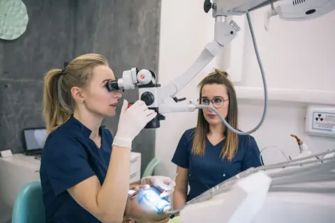 Dentystka z mikroskopem bada ząb pacjenta, by uniknąć chodzenia bez plomby. Pomaga jej asystentka.