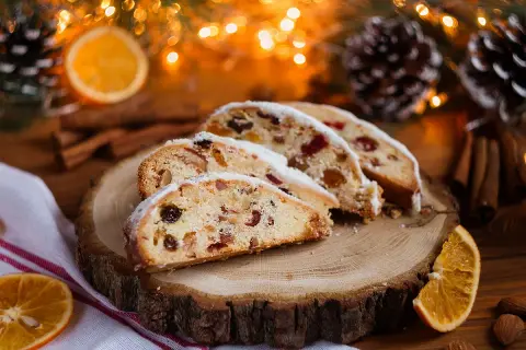 Tranches de stollen cake, un délice de Noël aux fruits secs et noix, saupoudré de sucre glace, sur une planche de bois, entouré de décorations festives.