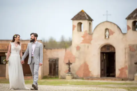 Un couple rayonnant, tout juste marié, se promène main dans la main devant une chapelle rustique, évoquant un mariage western.