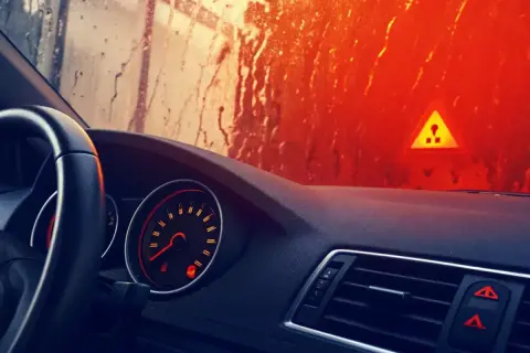 Tableau de bord d'une voiture, avec un voyant ABS allumé. Le soleil brille à travers un pare-brise mouillé, illuminant un panneau de danger.