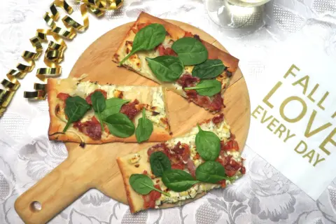 Prosciutto pizza med färsk spenat på en träskärbräda, omgiven av guldfärgad serpentin. En perfekt bit att "fall in love" med.