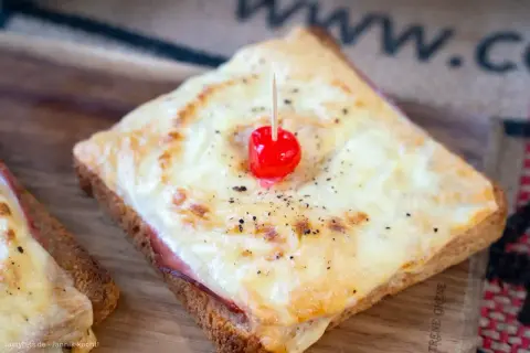 Ein klassisches Toast Hawaii mit geschmolzenem Käse, Schinken und einer Kirsche, serviert auf einem Holzbrett.